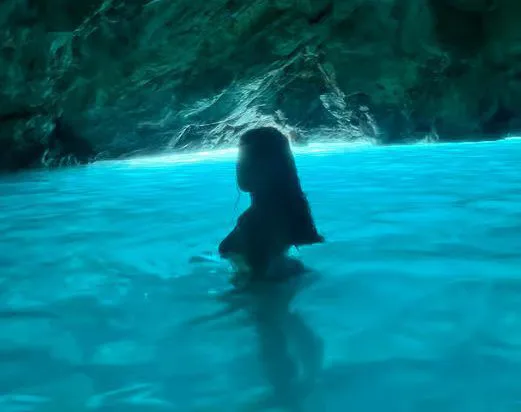 Woman wading in luminous blue water inside a coastal cave with rocky formations